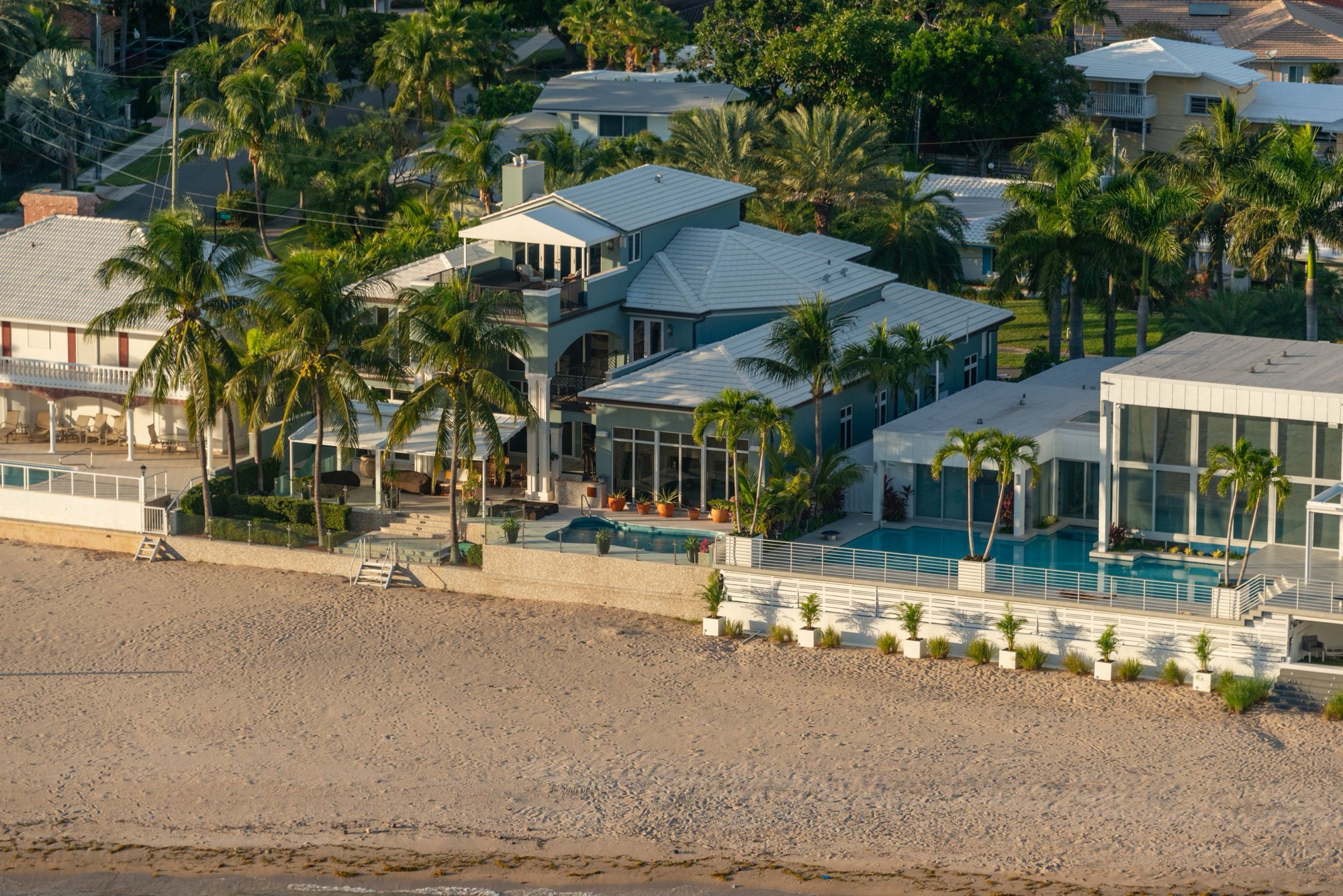 Beachfront houses with pools and palms photographed from the air in low golden light