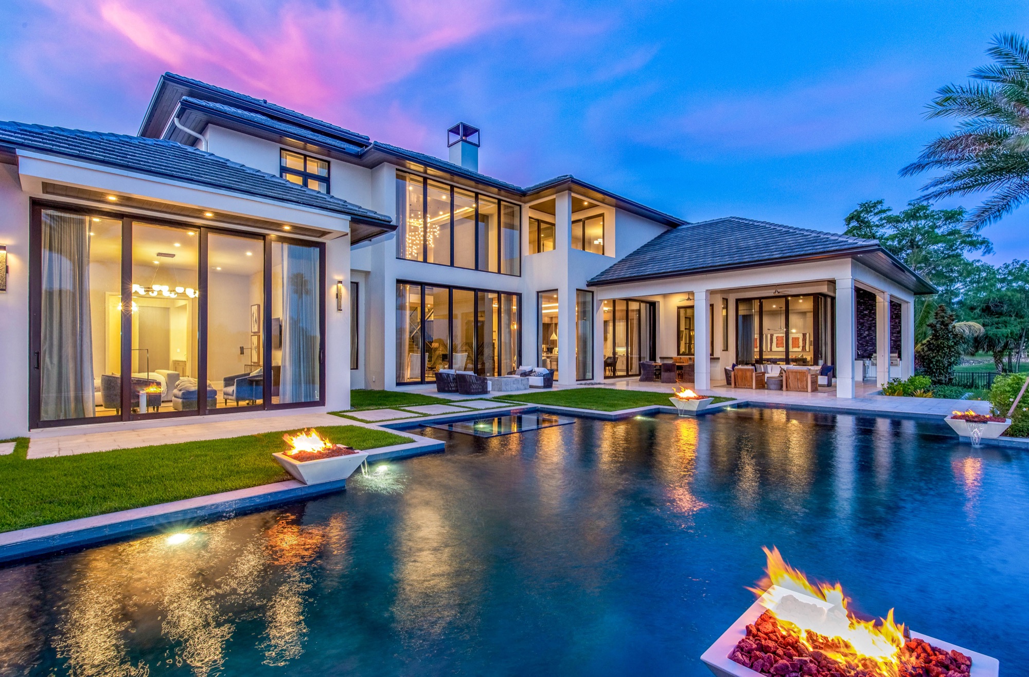 Modern luxury residence at dusk with a reflecting pool, lit fire bowls and glowing interiors